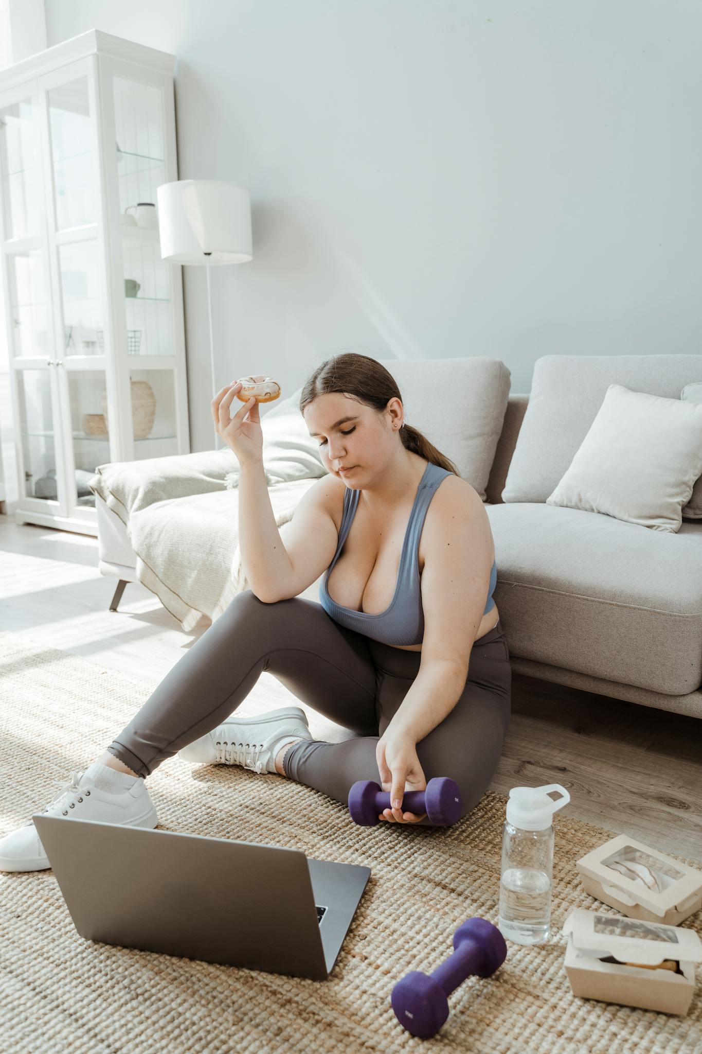 A woman in activewear sits with dumbbells and a donut beside a laptop, depicting balance.